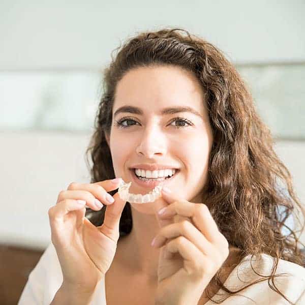 Woman with brown hair smiling as she shows a clear dental aligner held between her fingers.