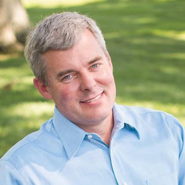 Man with short gray hair in a light blue shirt smiling outdoors in a sunny park setting.