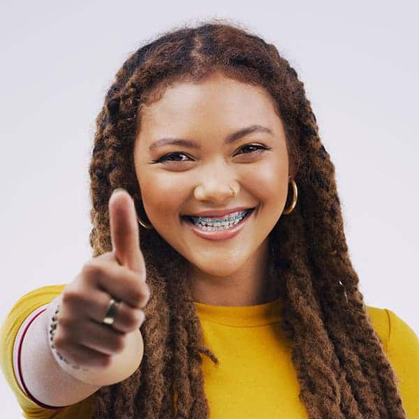 Smiling young woman with braces giving a thumbs-up, wearing a yellow top and hoop earrings.
