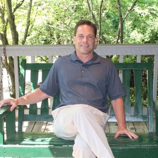 Man relaxing on a green wooden porch swing, smiling, wearing a blue polo shirt and light khaki pants, with trees in the background.